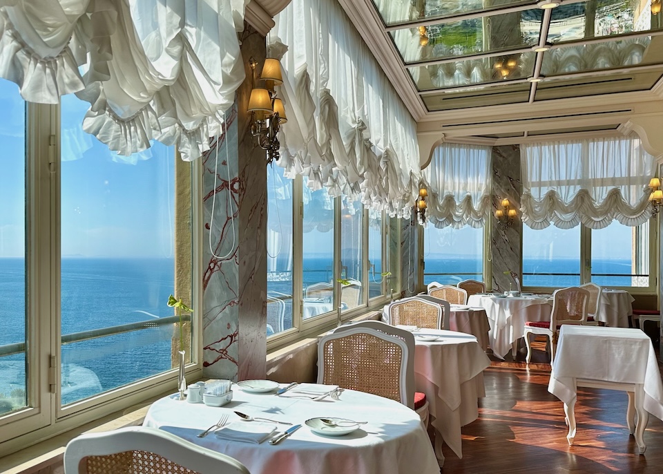 Wraparound sea-view windows with white, gathered curtains and white tablecloths in the dining room of Excelsior Grand Vittoria hotel in Sorrento