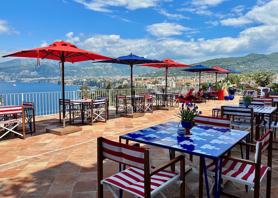 Bold red, white, and blue furniture and umbrellas on a terracotta-floor terrace facing the sea and city at Maison la Minervetta hotel in Sorrento