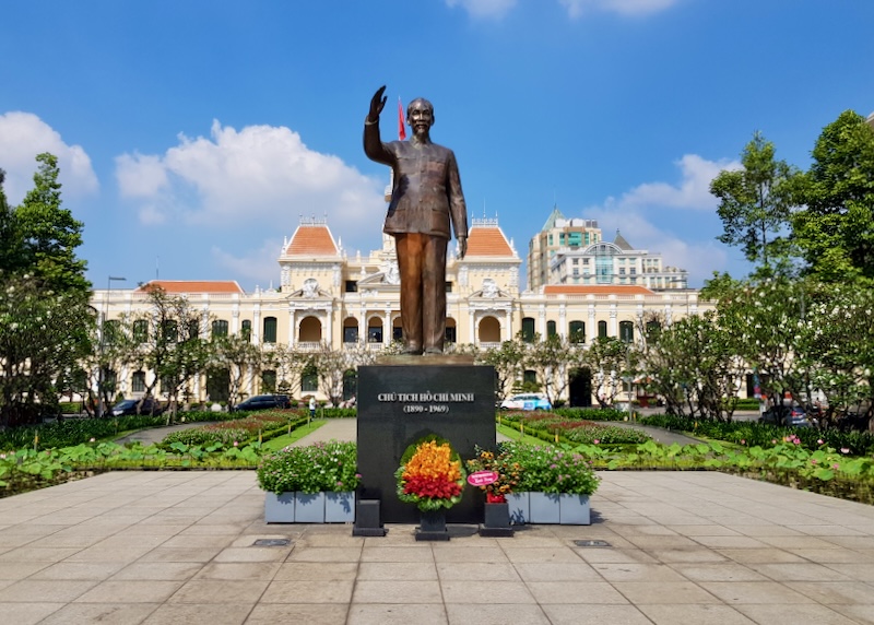 A bronze statue of the man, Ho Chi Minh, stands tall with his right arm raised as if to say hello.