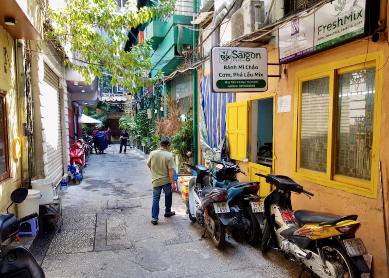 A man walks down an alley with bright green and yellow painted shops as scooters rest upon their walls.