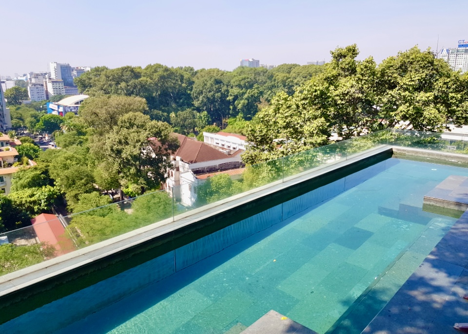 A narrow rectangle pool sits a top a hotel overlooking large green trees.