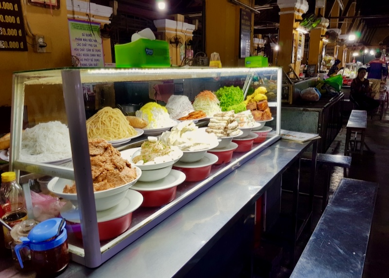 Dishes of food sit behind glass at a food stall at a Market.