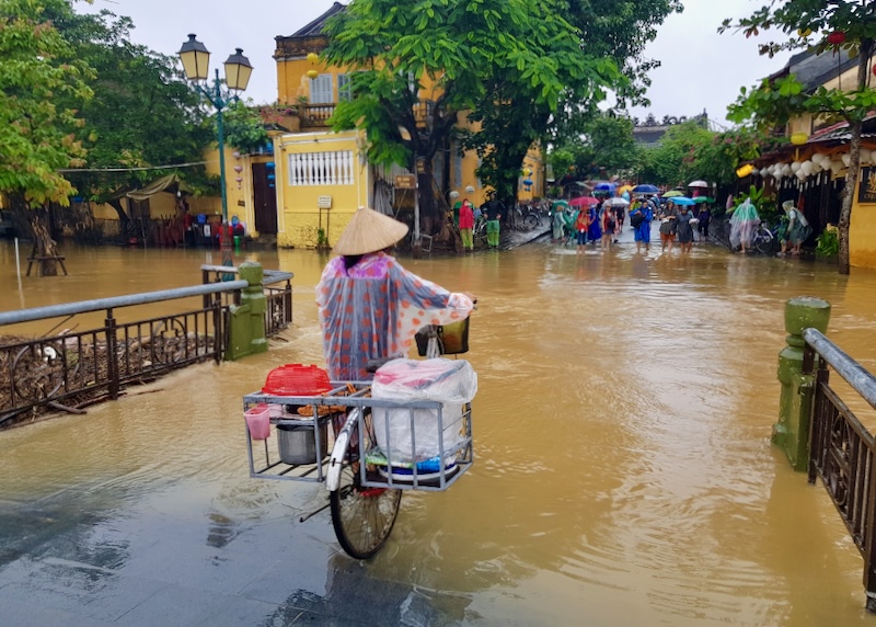 A woman on a bike rides into yellow-brown flood waters.