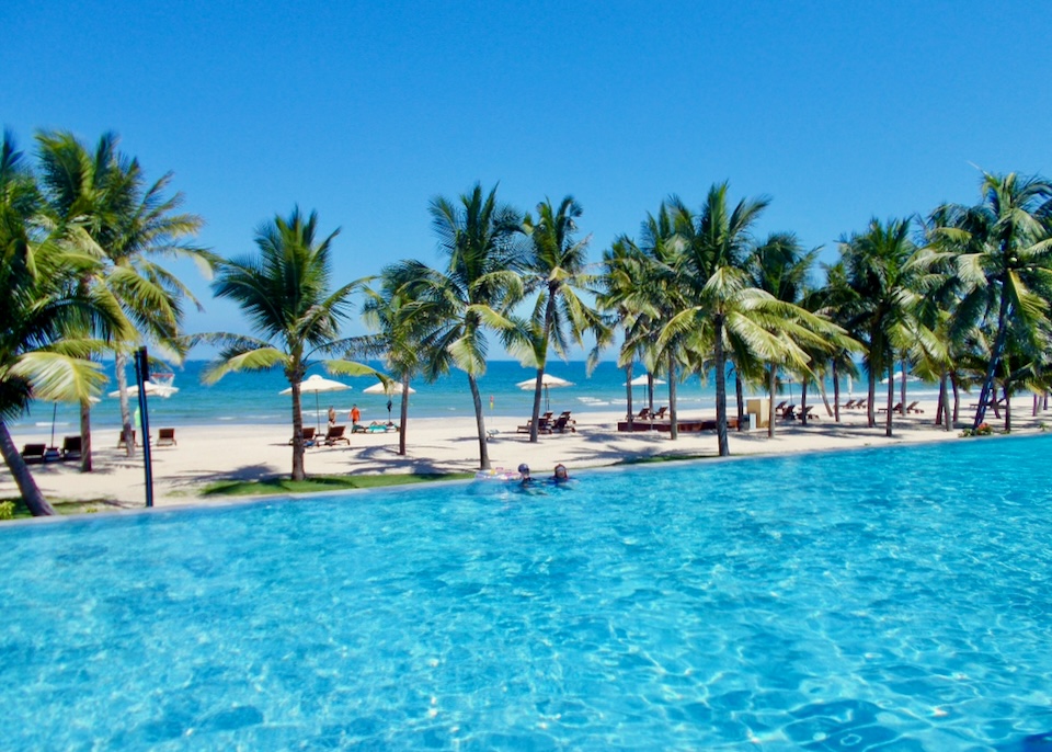 A light blue pool sits next to the beach separated by lush palm trees.