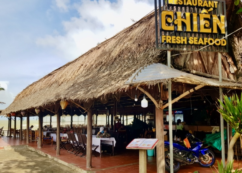 A thatched-roof open-air seafood restaurant sits next to the sand and ocean.