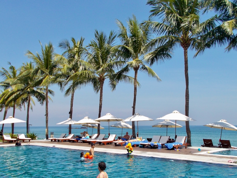 Families swim a a pool lined with palm trees next to the ocean.