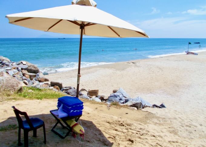 A tan sand beach with an umbrella shading a chair on a hill.