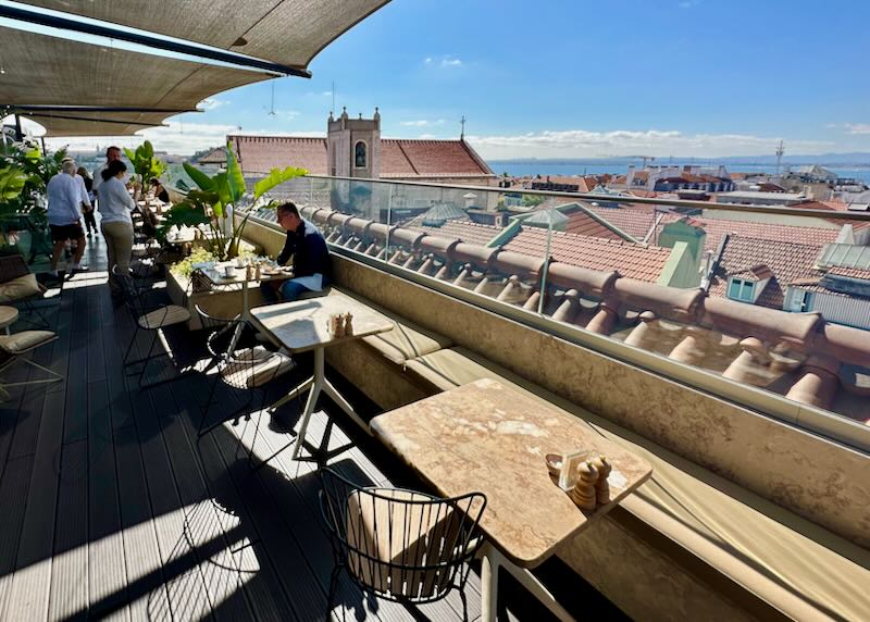 A man sits at a rooftop cafe table, overlooking the rooftops of Lisbon