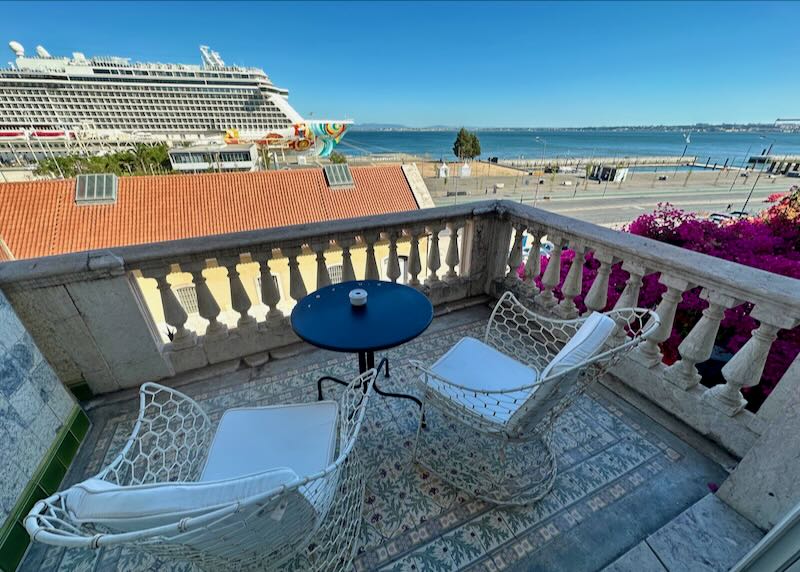 Hotel balcony with two chairs and a cafe table overlooking a seaport with a cruise ship at dock.