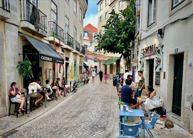 People sit at small outdoor cafe tables in a narrow pedestrian street lined with cafes