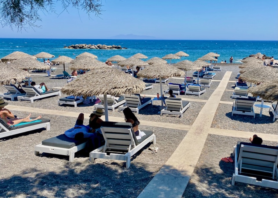 On the black beach with blue sea lined with rows of sunbeds and thatched umbrellas in Santorini