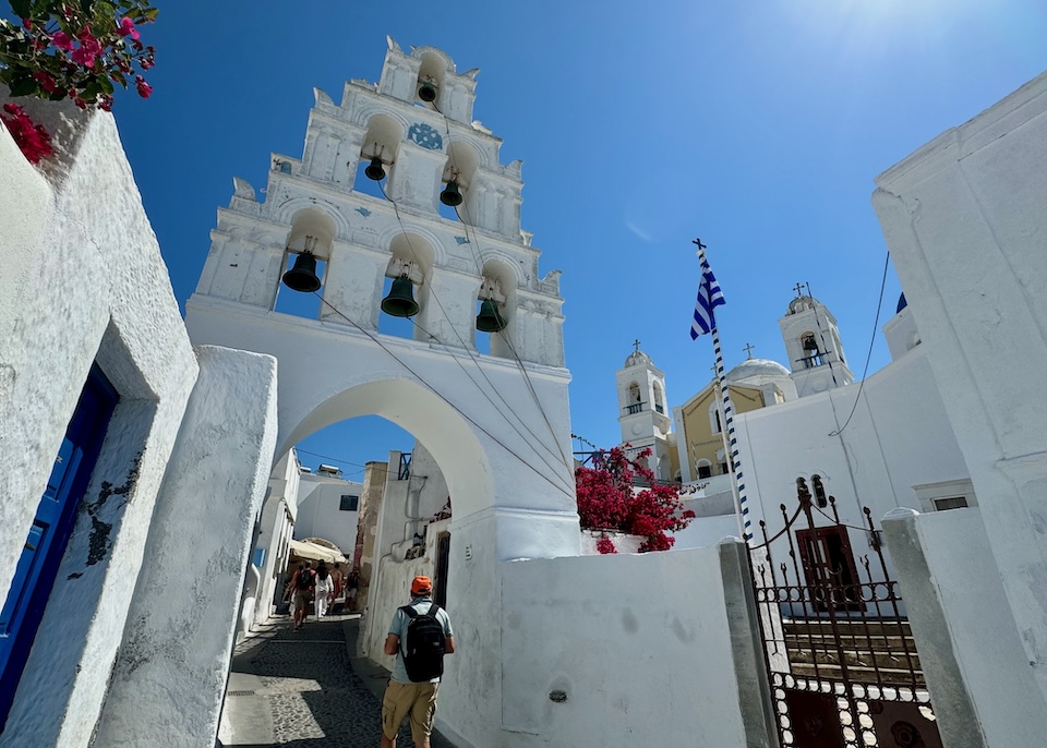 A centuries-old bell tower crosses a pedestrian lane in front of a church in a traditional village in Santorini