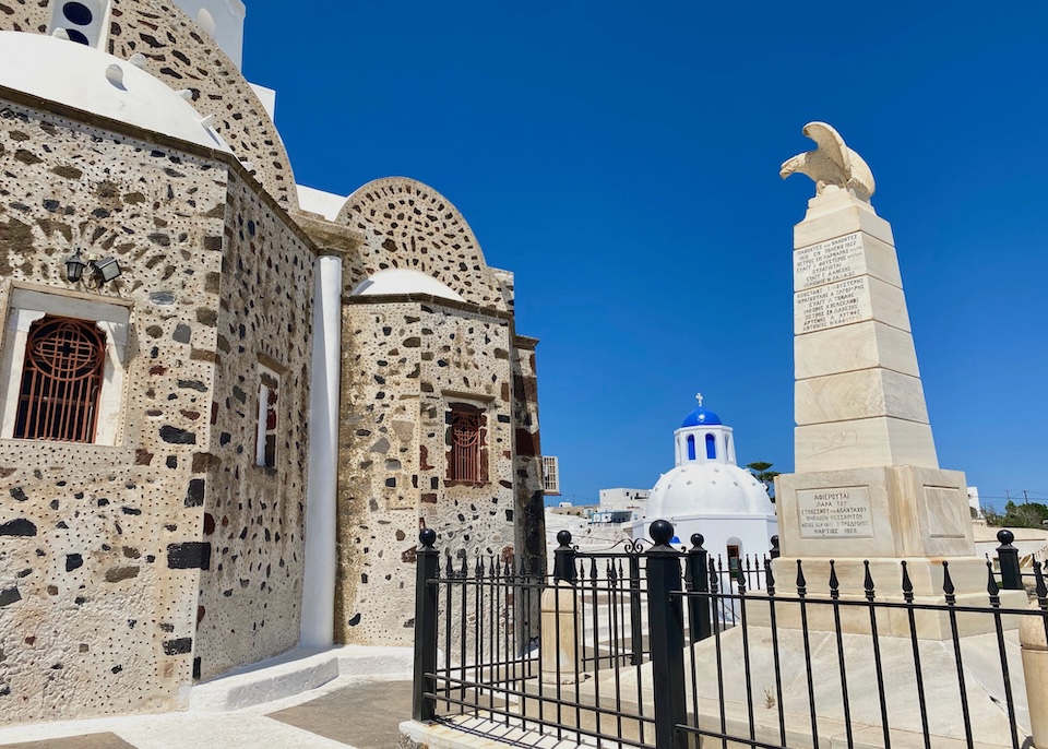 A stone-built church and eagle-topped statue in the foreground and the white and blue-dome of another church in the background in a village in Santorini