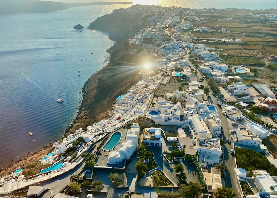 Aerial view over Oia village with white hotels, blue pools, and boats on the caldera in Santorini
