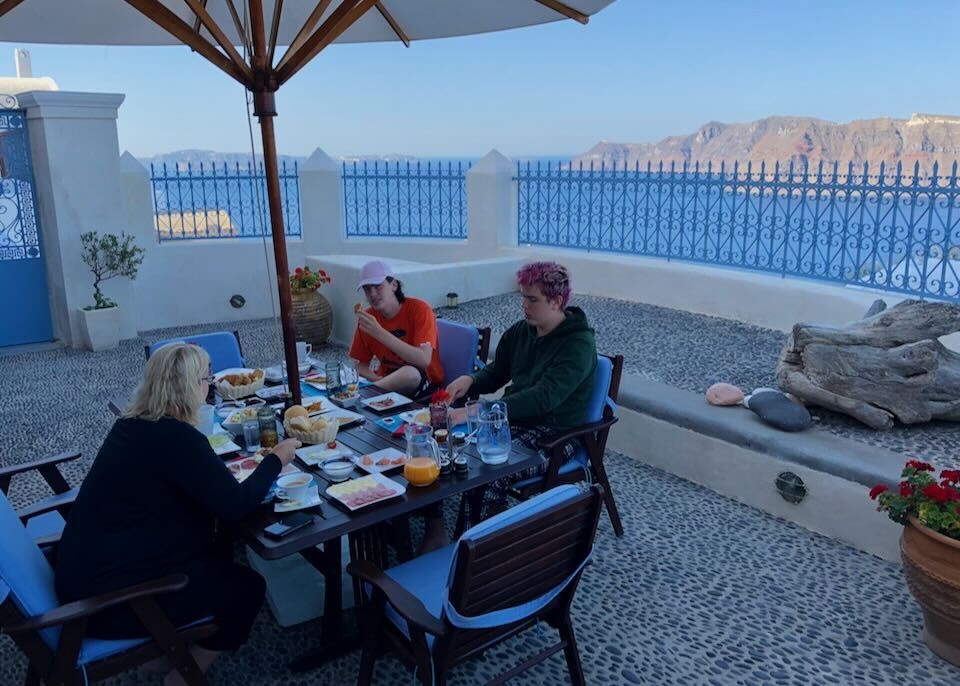 A woman and two teenagers have breakfast on a pebbled terrace with a view to the caldera and sea in Santorini, Greece