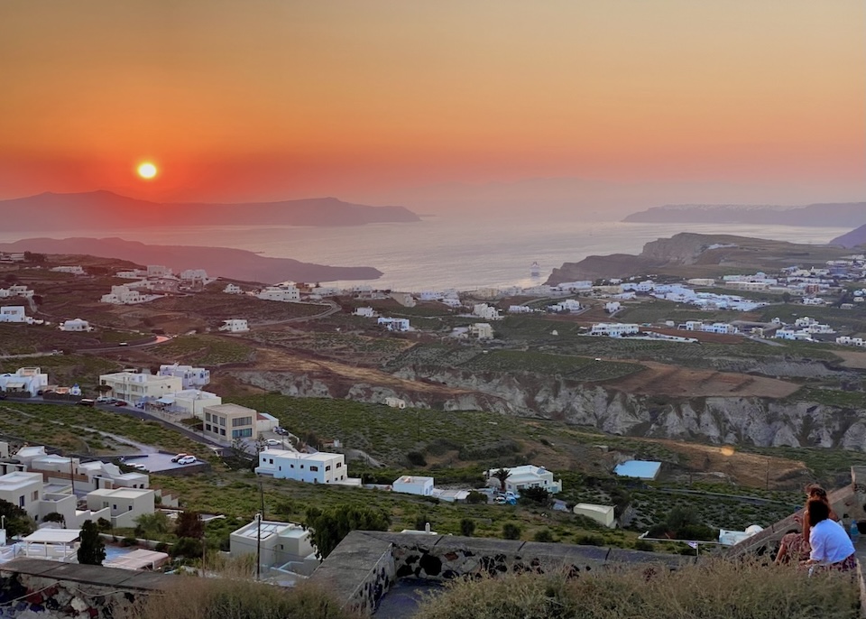 A vibrant red-orange sunset over the sea with islands in the background and vineyards and houses in the foreground.