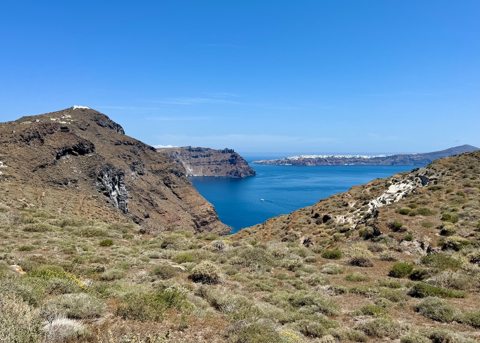 The wilderness of Thirassia, covered in low-lying shrubs with cliffs, a the caldera spreading below, and Oia village in Santorini in the distance