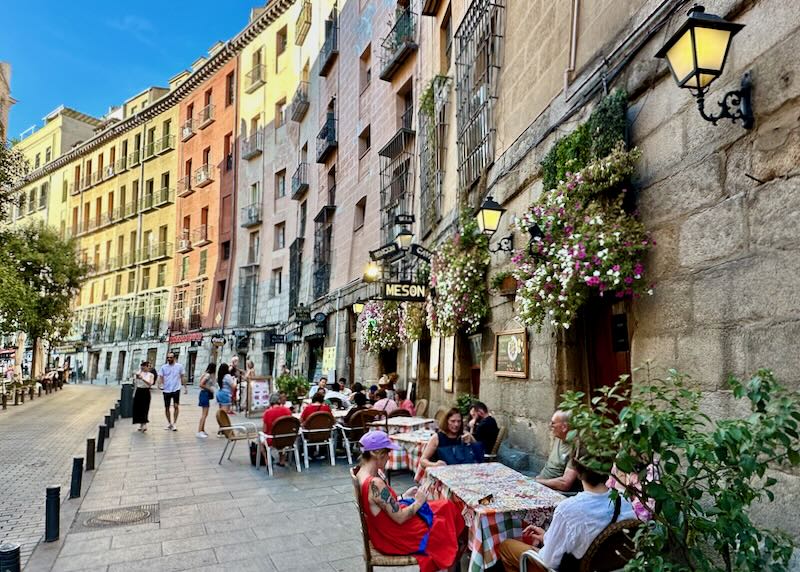 Cafe tables along a sidewalk lined with colorful Spanish buildings.