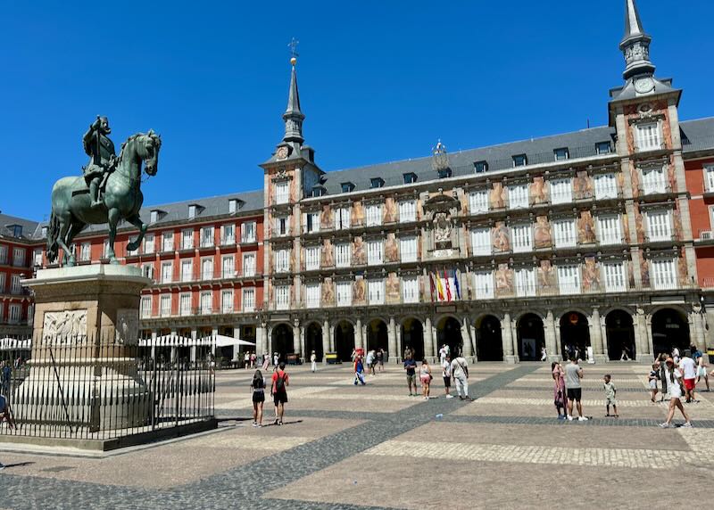 Large ornate buildings lining the Plaza Mayor in Madrid, with many pedestrians walking past.