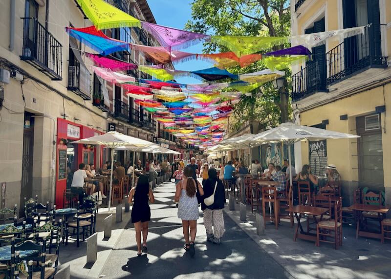 Narrow street lined with shops and overhung with colorful scarves, with many pedestrians walking on the sidewalks