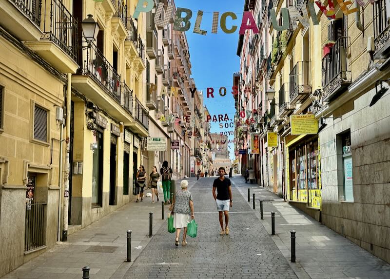 A man walks down a narrow street that is overhung with colorful letters