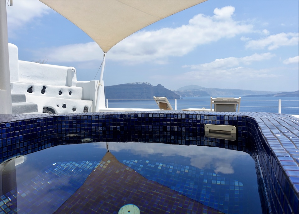 A dark blue tiled jacuzzi under a sail clothe canopy with a south-facing view over the caldera toward Skaros Rock at Ambition Suites in Oia, Santorini.