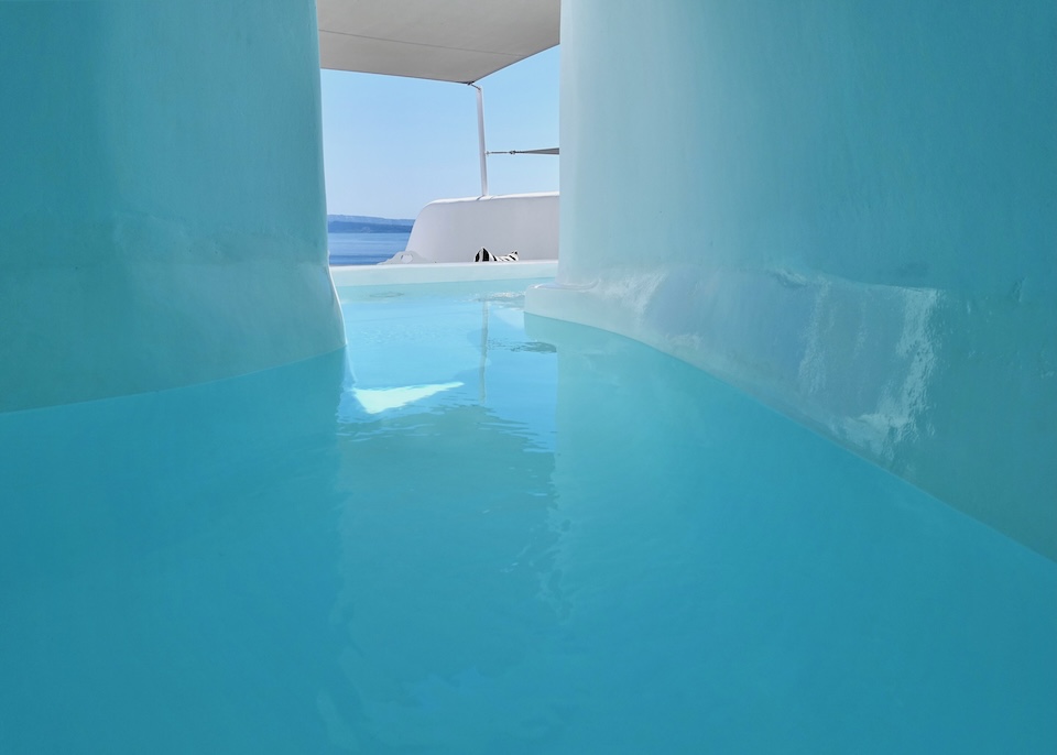 A long, winding, cave pool with blue reflected on the walls and a view toward the caldera and a whitewashed terrace at Canaves Ena hotel in Oia, Santorini