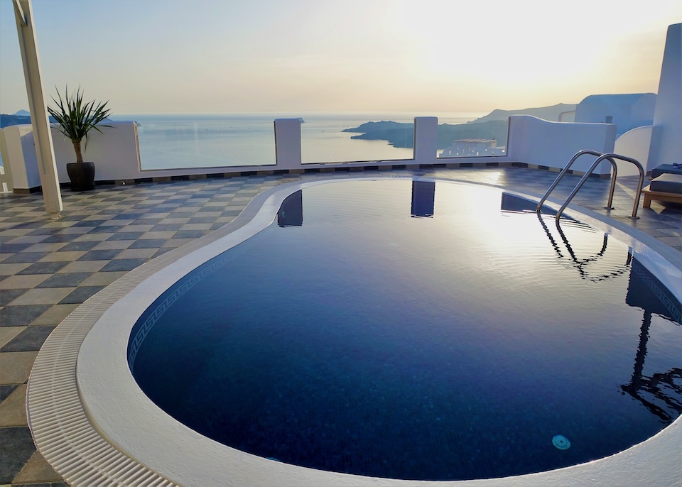 A sunken, kidney-shaped, infinity pool on a checkerboard terrace reflects the light near sunset on the caldera's edge at Celestia Grand in Mesaria, Santorini.