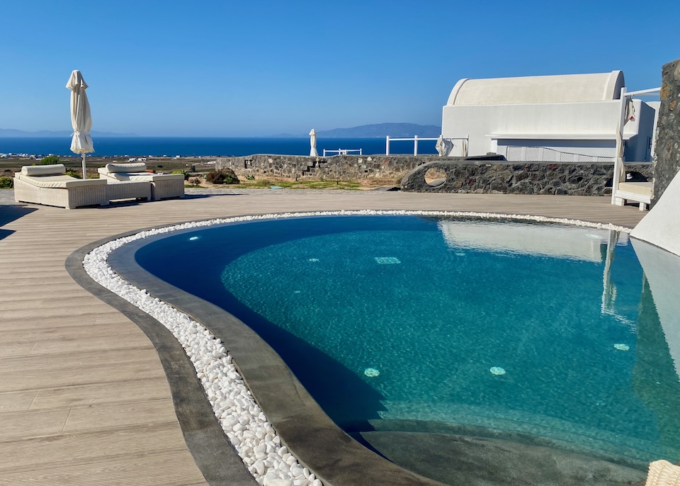 A freeform infinity edge pool on a wooden deck with sunbeds and a Bali bed facing east toward the sea at Danae Suites in Finikia, Santorini.