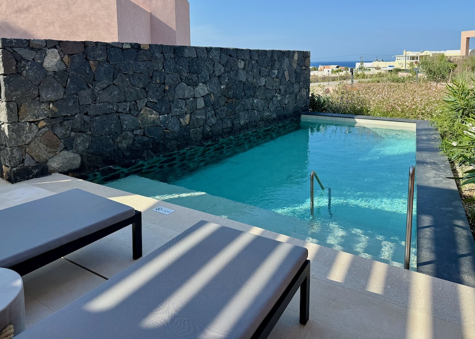 A private pool facing the garden and sea in the distance with a lava rock wall separating the pool from it's neighbors at Domes Novos Resort near Oia, Santorini.