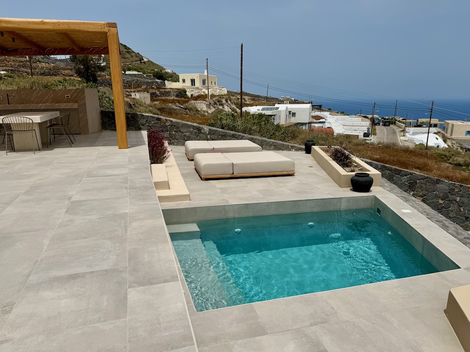 A private plunge pool on a sunny terrace with sunbeds and a dining area plus the sea in the distance at Ethos Vegan Retreat in Imerovigli, Santorini.