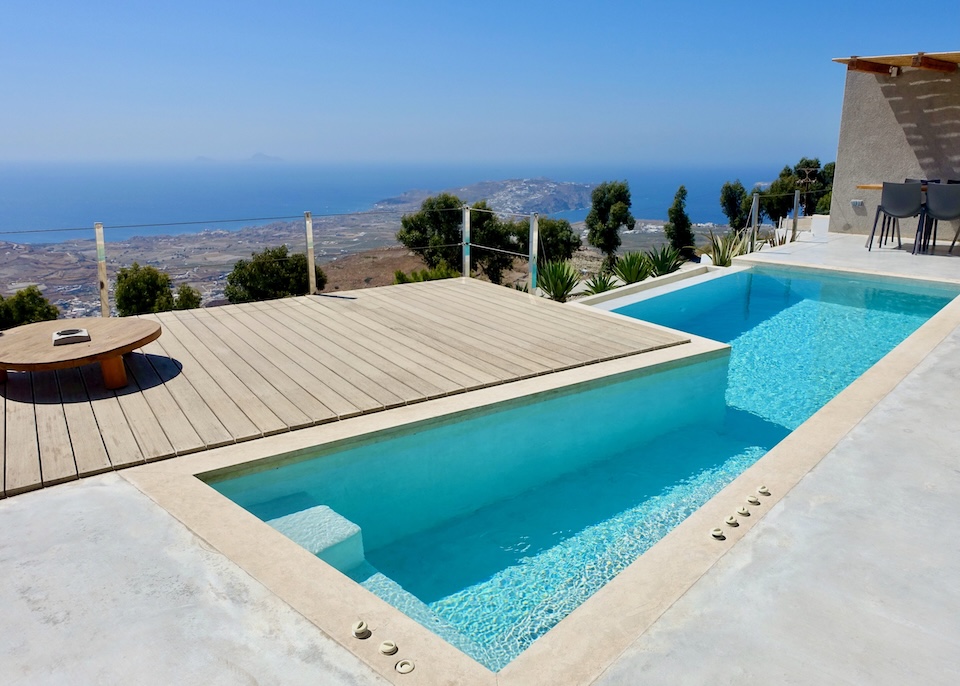 An angluar infinity pool with a large wooden deck and a pergola-covered dining area and panoramic views from Santorini Heights at Imerovigli, Santorini.