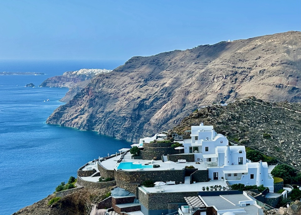 Cycladic-style arhitecture with barrel roofs, boxy walls, and a pool fronted by a sun terrace above the caldera with Oia village in the distance at Aenaon Villas in Imerovigli, Santorini.