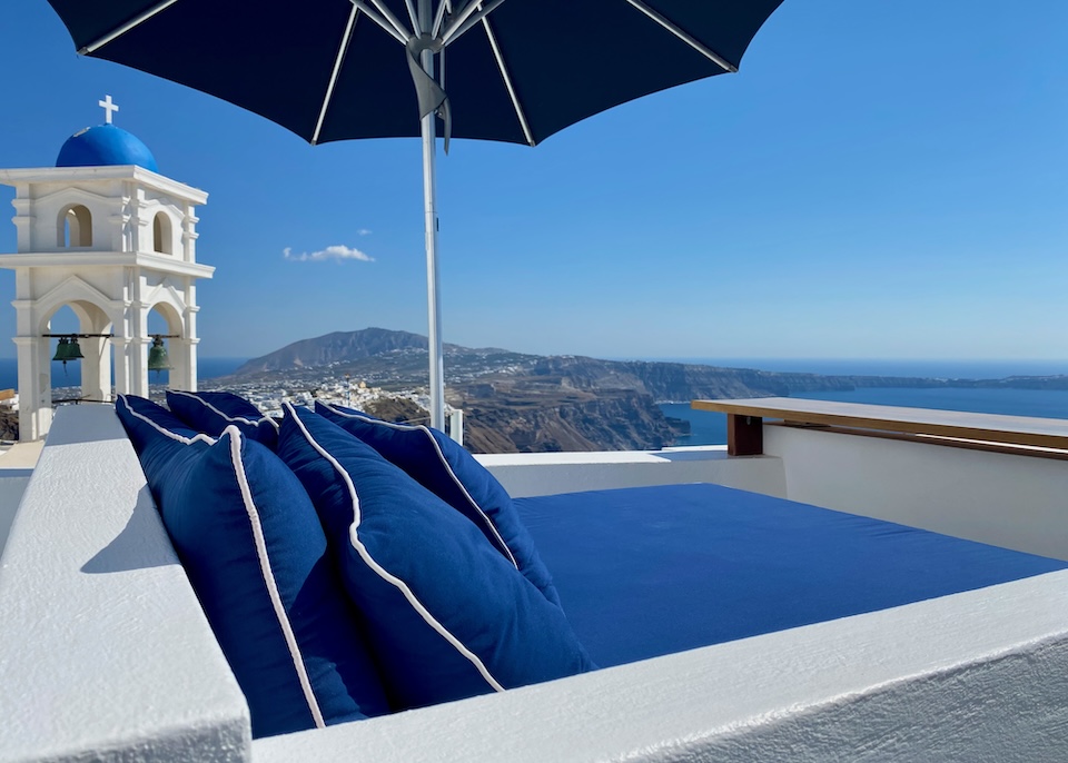 A double-sized sunbed shaded by an umbrella sits beneath a blue-capped bell tower and above the caldera at Altana Heritage Suites in Imerovigli, Santorini.