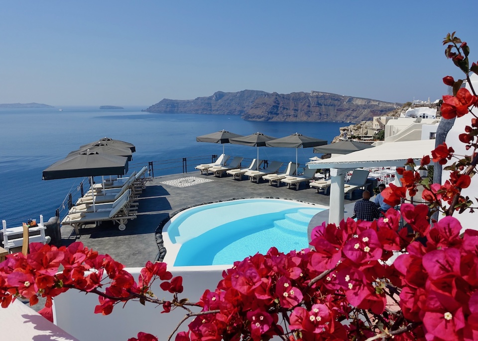 Magenta bougainvillea flowers frame a pool with a sun terrace jutting out over the caldera toward Thirassia Island at Andronis Boutique Hotel in Oia Santorini.