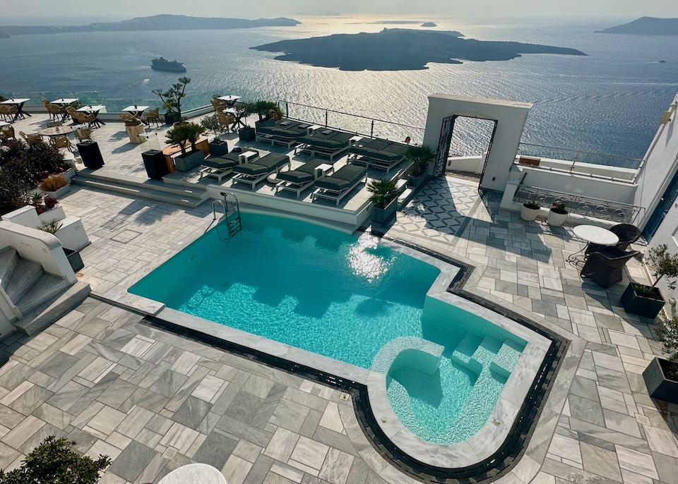A freeform pool with attached jacuzzi sits on a terrace with sunbeds and a view of the caldera and volcanoes at Anteliz Suites in Firostefani, Santorini.