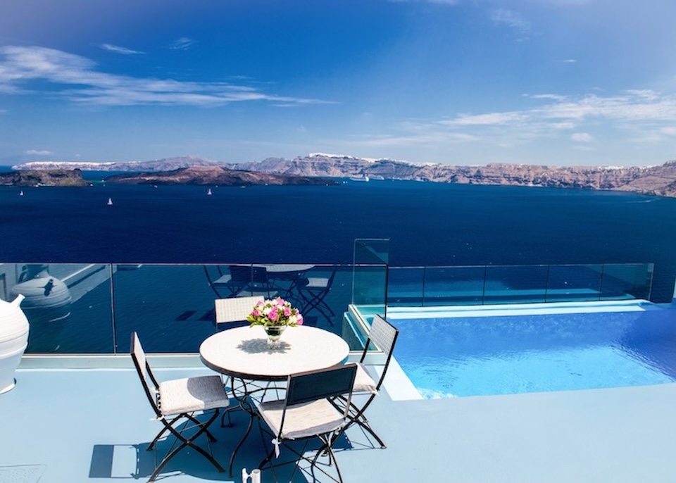 An infinity edge pool appears to hang over the caldera next to a dining table on a glass-in terrace at Astarte Suites in Akrotiri, Santorini.