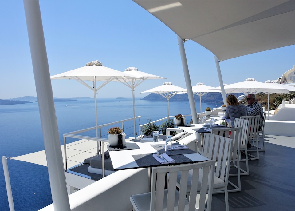 A dining terrace with white umbrellas over a row of tables seems to float above the caldera at Canaves Ena hotel in Oia, Santorini.