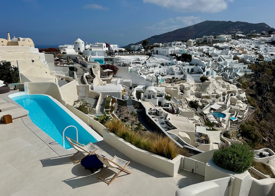 A sun terrace purched atop the cliff with two lounge chairs, an infinity pool and a view of Oia village at Canaves Suites in Santorini