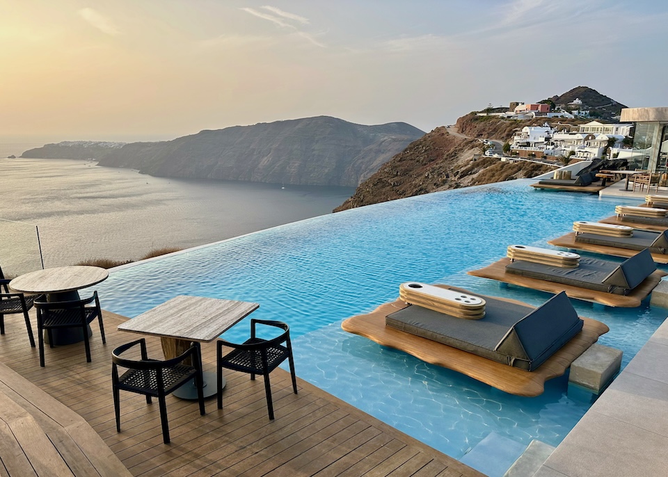 A dining terrace next to a row of floating sunbeds in an infinity pool with a view over the caldera and toward the cliffs and Oia village in the distance at Cavo Tagoo hotel in Imerovigli, Santorini.