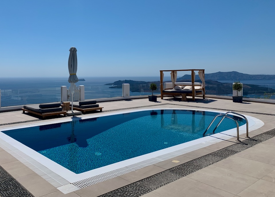 A infinity pool in the middle of a terrace with sunbeds and a Bali bed overlooking the caldera at Celestia Grand hotel in Mesaria, Santorini.