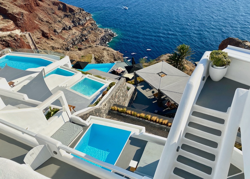 Different-sized private pools next to a staircase spread on terraces down the side of the caldera with a few small boats in the water below at Charisma Suites in Oia, Santorini.