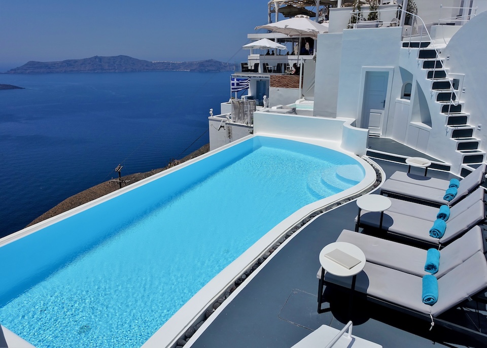 An infinity pool with a row of sunbeds behind, a stairway leading up to hotel rooms, and a view over the caldera at Cosmopolitan Suites in Fira, Santorini.