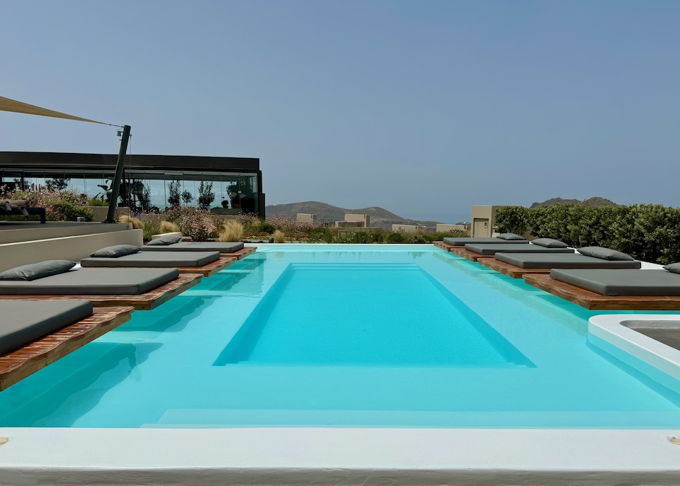 A rectangular infinity pool with double-sized floating sunbeds on the long sides and a restaurant in the back at Divine Cave Experience in Imerovigli, Santorini.