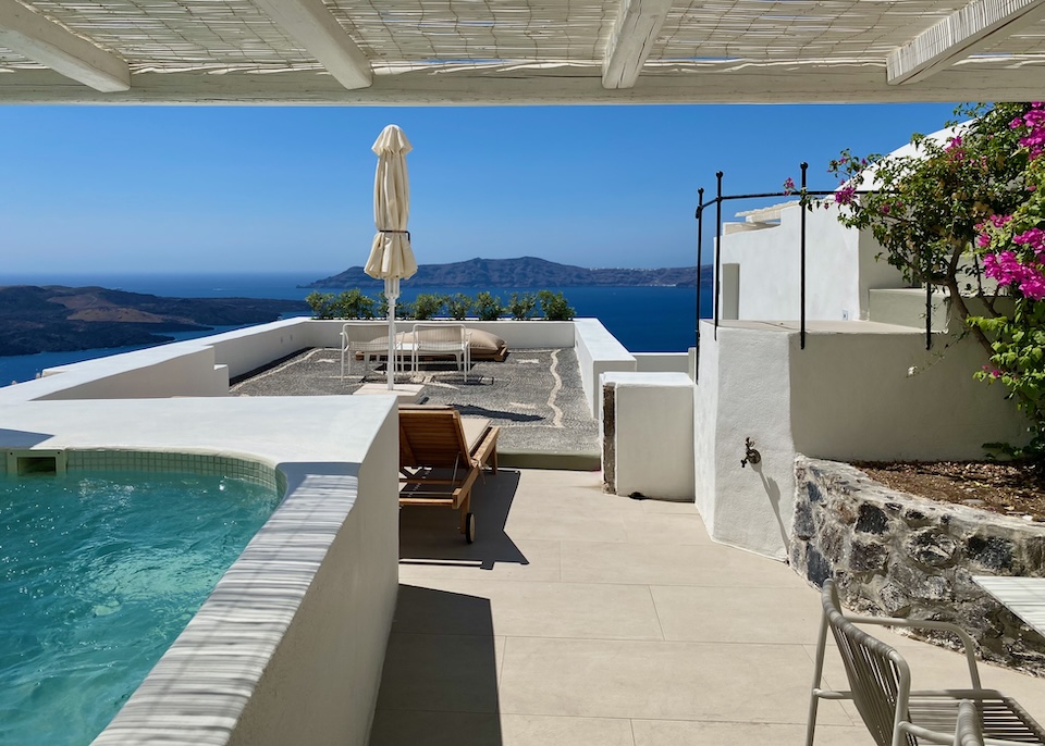 A plunge pool and dining table are shaded by a pergola, while a sunbed and lounge area sit under the sun with a view of the caldera at Enigma Suites in Fira, Santorini.