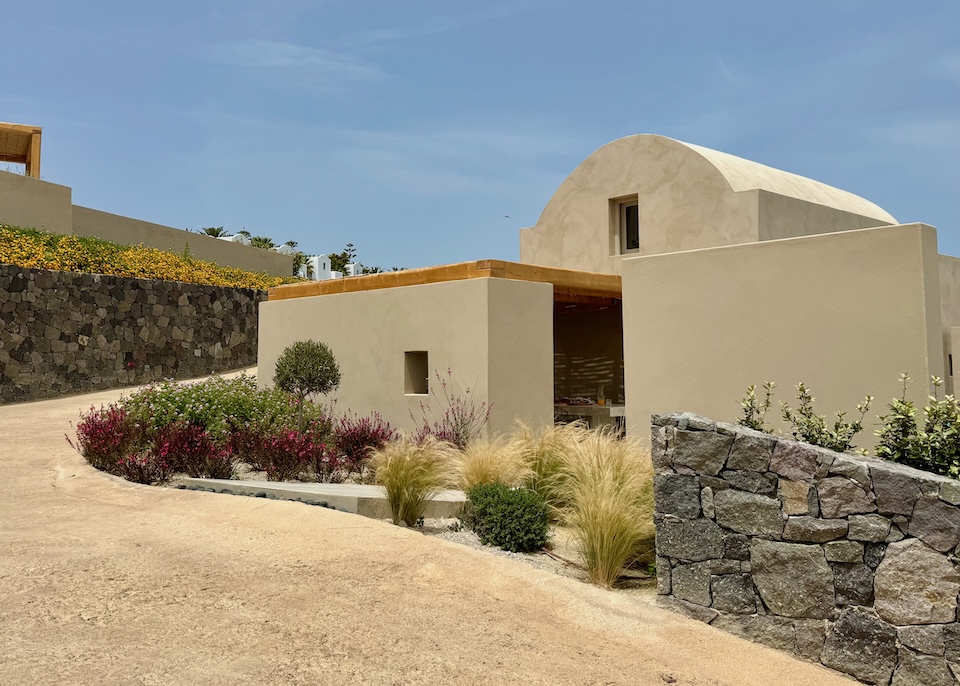 The exterior of a villa featuring modern, Cycladic architecture, lava rock walls, and a garden at Ethos Vegan Retreat in Imerovigli, Santorini.