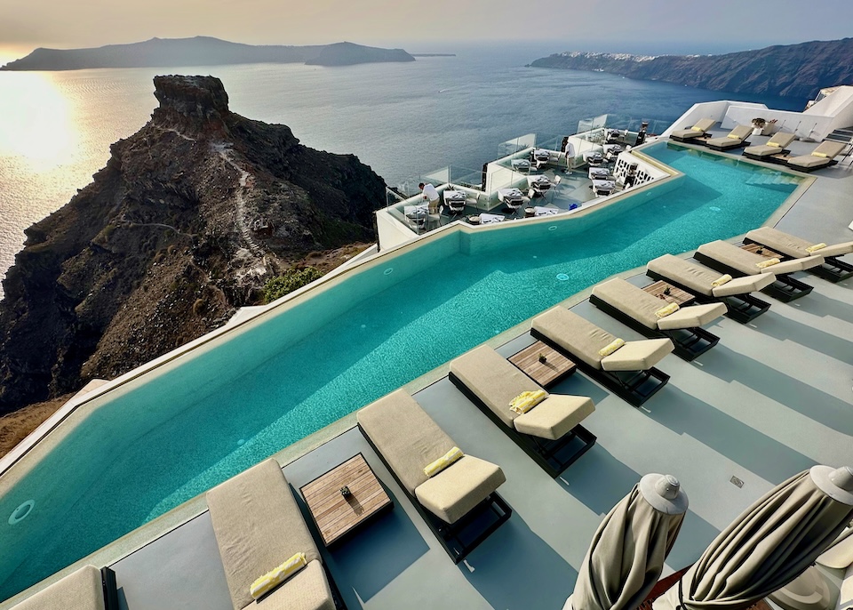 A long infinity pool with lined with sunbeds overlooks the caldera and Skaros Rock in Imerovigli, Santorini.