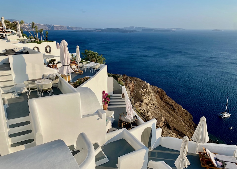 A series of furnished terraces, balconies, and stairways overlook the caldera and a catamaran at Ikies hotel in Oia, Santorini.