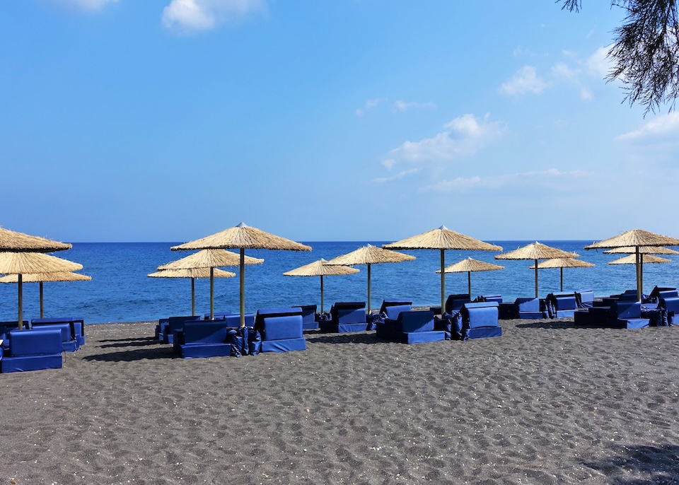 Three rows of plush, blue sunbeds under golden straw umbrellas sit on a black sand beach facing the Aegean Sea at Istoria hotel on Perivolos Beach in Santorini.