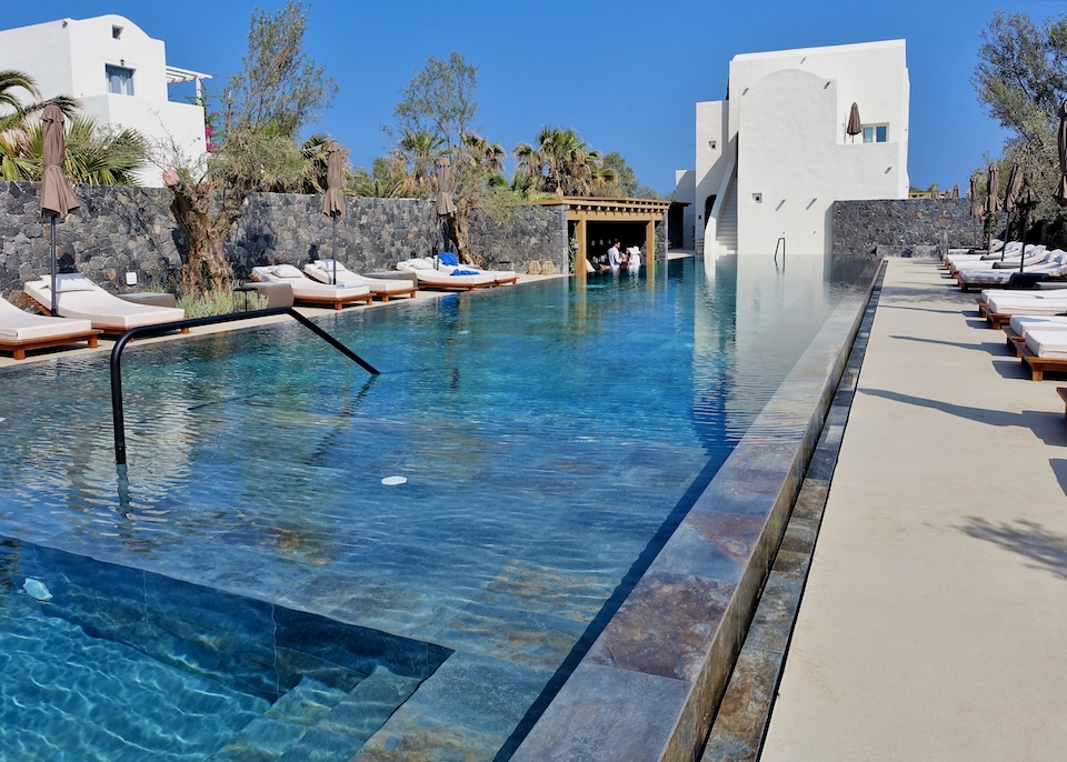 A long, stone-built infinity pool with sunbeds on both sides facing a Cycladic-style building at Istoria Hotel in Perivolos, Santorini.